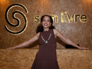 Bruna Black assina com Som Livre (Crédito: André Rola) Mulher sorridente em um ambiente moderno, posando em frente ao letreiro 'som livre', destacando sua expressão confiante e estilo elegante.