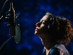 Marisa Monte em estúdio de gravação, usando fones de ouvido e com expressão intensa, capturada contra fundo azul desfocado.