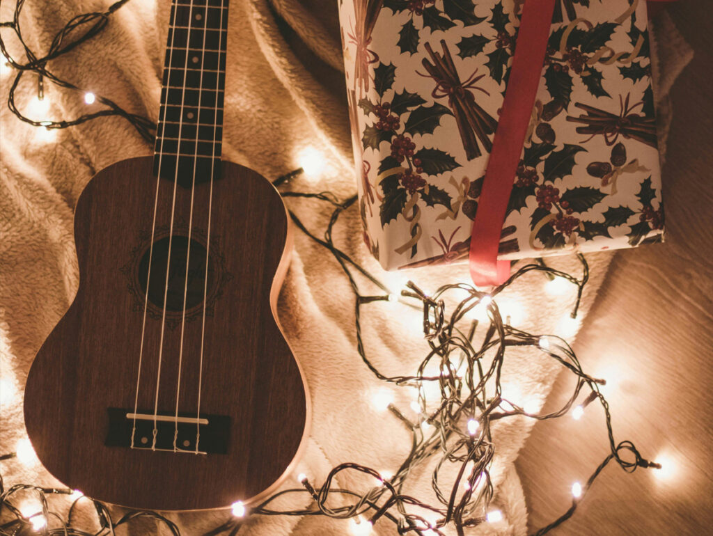 Um ukulele em cima de uma manta, cercado por luzes de Natal e um presente decorado com folhas de azevinho e fita vermelha, criando um ambiente natalino aconchegante.
	
