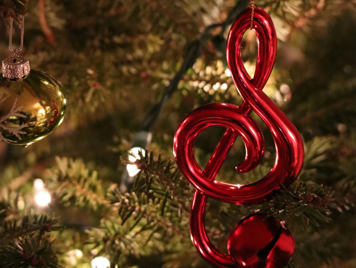 A imagem mostra um ornamento de Natal em forma de clave de sol vermelha pendurado em uma árvore decorada, simbolizando o espírito festivo e musical.