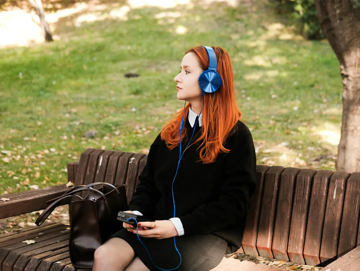 Uma jovem com cabelos ruivos usa fones de ouvido azuis enquanto relaxa em um banco em um parque, segurando um dispositivo de música. A imagem captura um momento de tranquilidade e estilo. Ideal para quem busca inspiração em moda jovem e cultura musical.