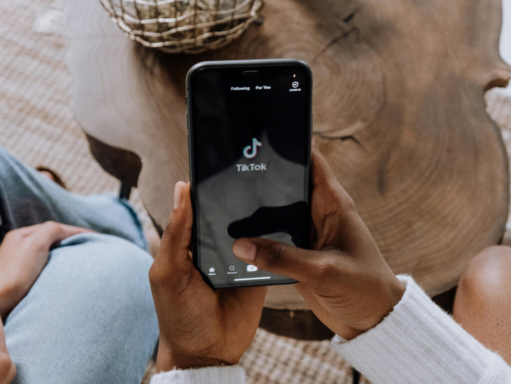 Usuário acessando o aplicativo TikTok em um smartphone, destacando a interface do app com a logo do TikTok. O cenário apresenta um ambiente confortável e aconchegante. Estudo da Luminate avalia o impacto do app.